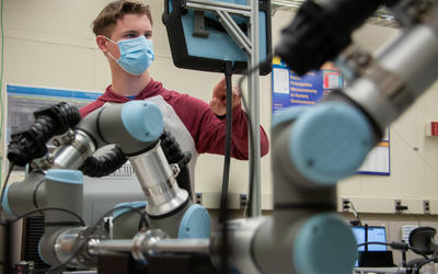 A man wearing a face mask touches a computer screen in a lab with robotic arms in the foreground.