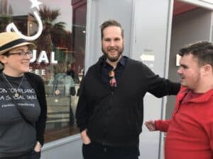 Three white autistic people. Two adults: Sara Luterman and John Marble, and one teen, Leo Rosa, hanging out in front of a coffee shop.