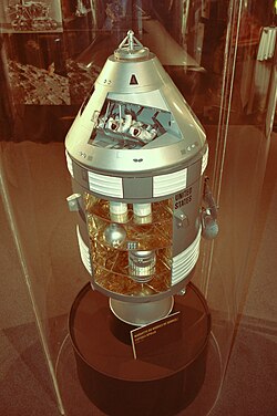 The model, displayed in a cylindrical glass case, has cutaways showing the three astronauts in the CM and the contents of one of the SM segments. It is labelled in French.